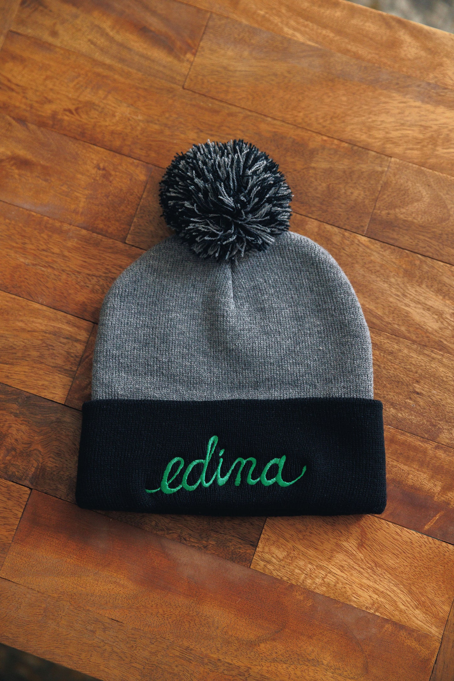 Gray beanie with black band and green 'edina' text on a wooden surface