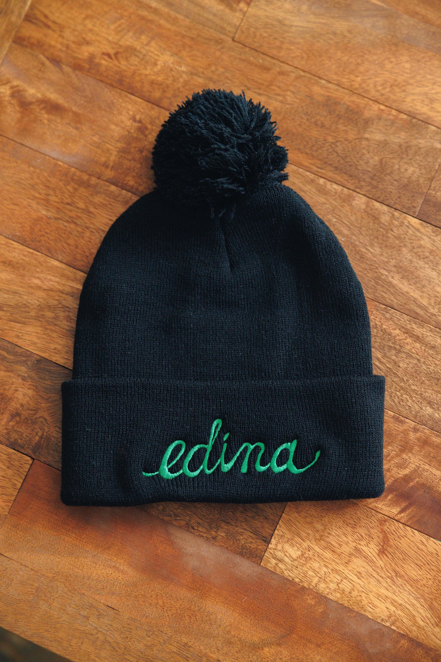 Black beanie with a pom-pom and 'edina' embroidery on a wooden surface