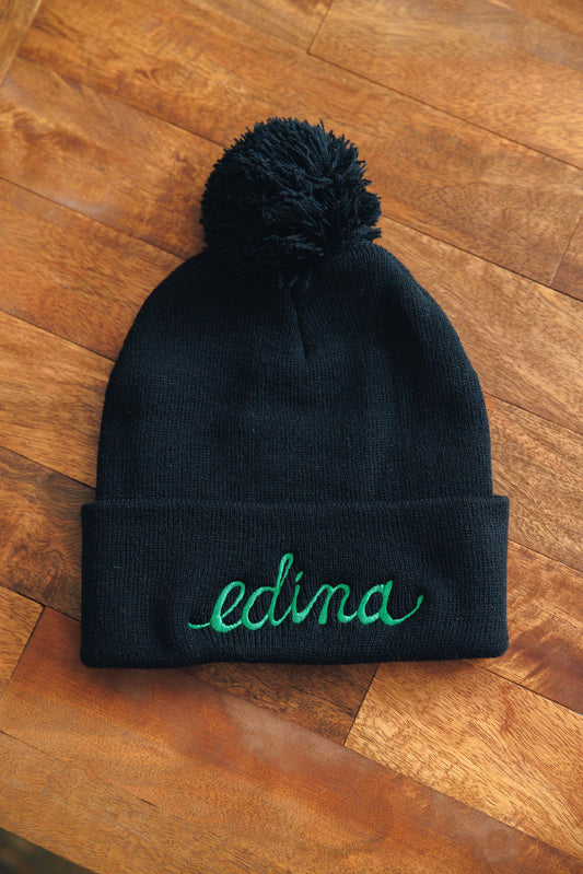Black beanie with a pom-pom and 'edina' embroidery on a wooden surface