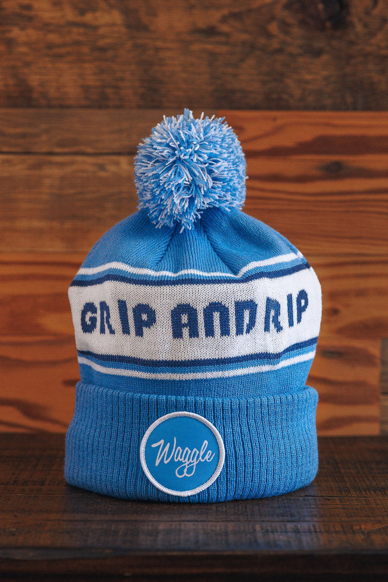 Blue beanie with pom-pom and 'Grip And Rip' text on a wooden background