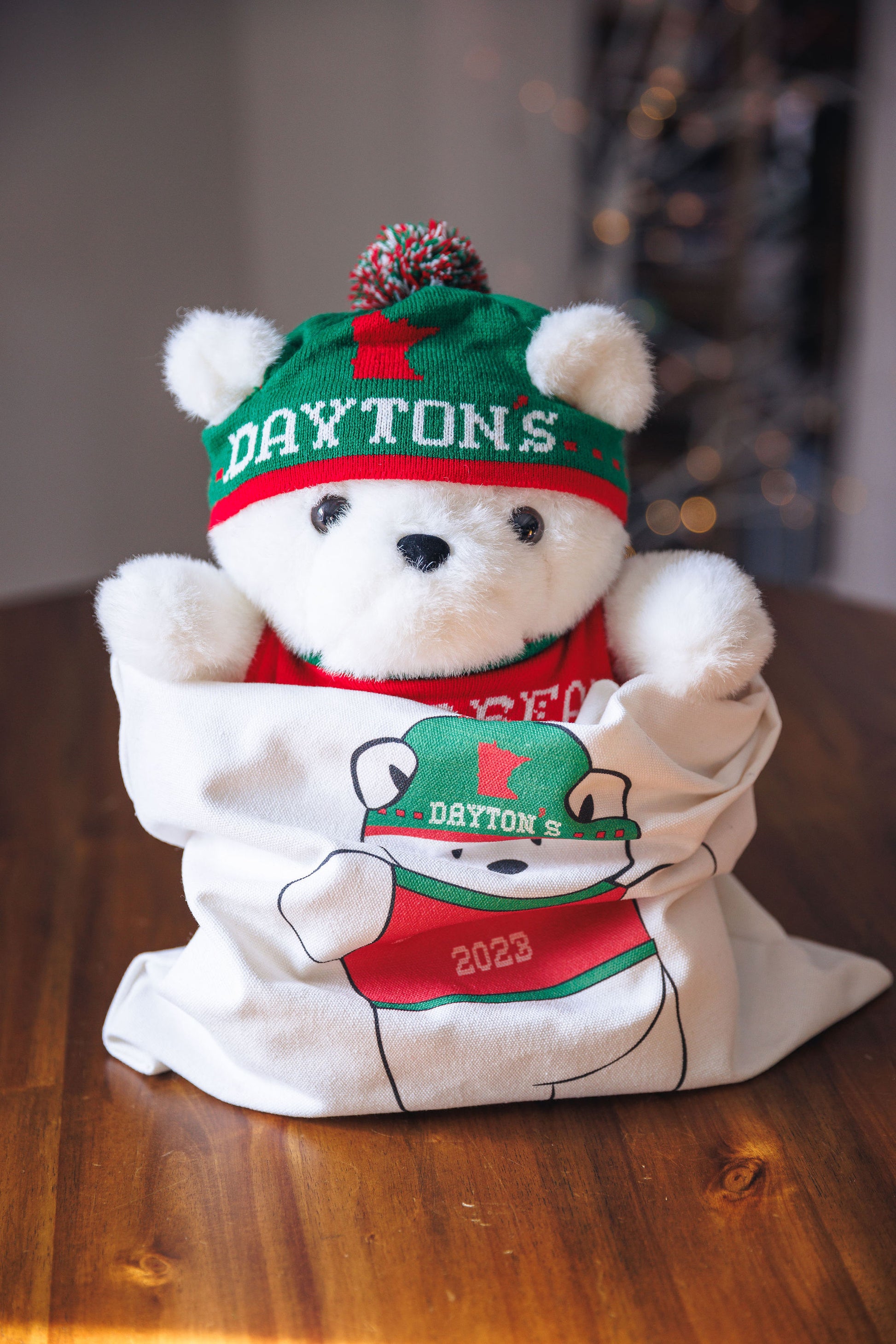 White teddy bear wearing a green and red 'Daytons' hat and shirt on a wooden surface.