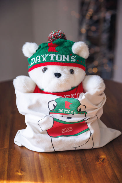 White teddy bear wearing a green and red 'Daytons' hat and shirt on a wooden surface.