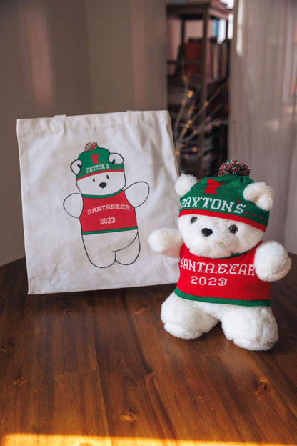 White teddy bear in a red sweater and green hat next to a tote bag with a similar design on a wooden surface.