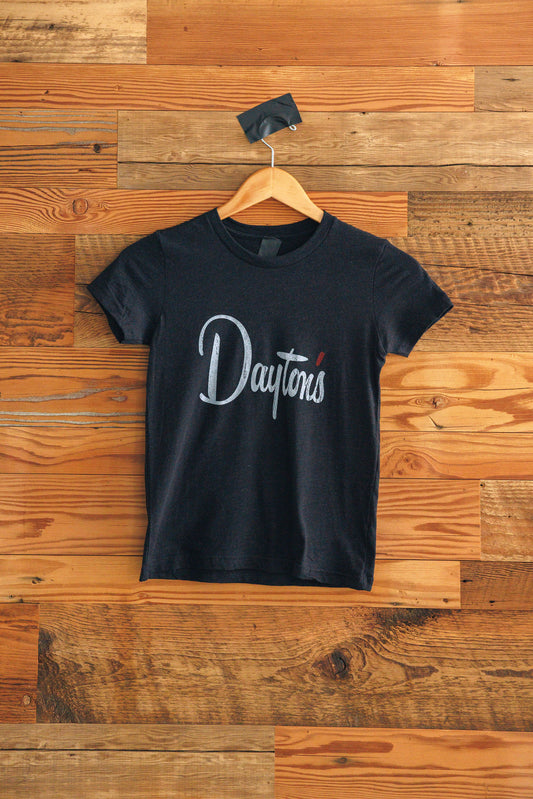 Black t-shirt with 'Dayton' printed on it, hanging against a wooden wall.
