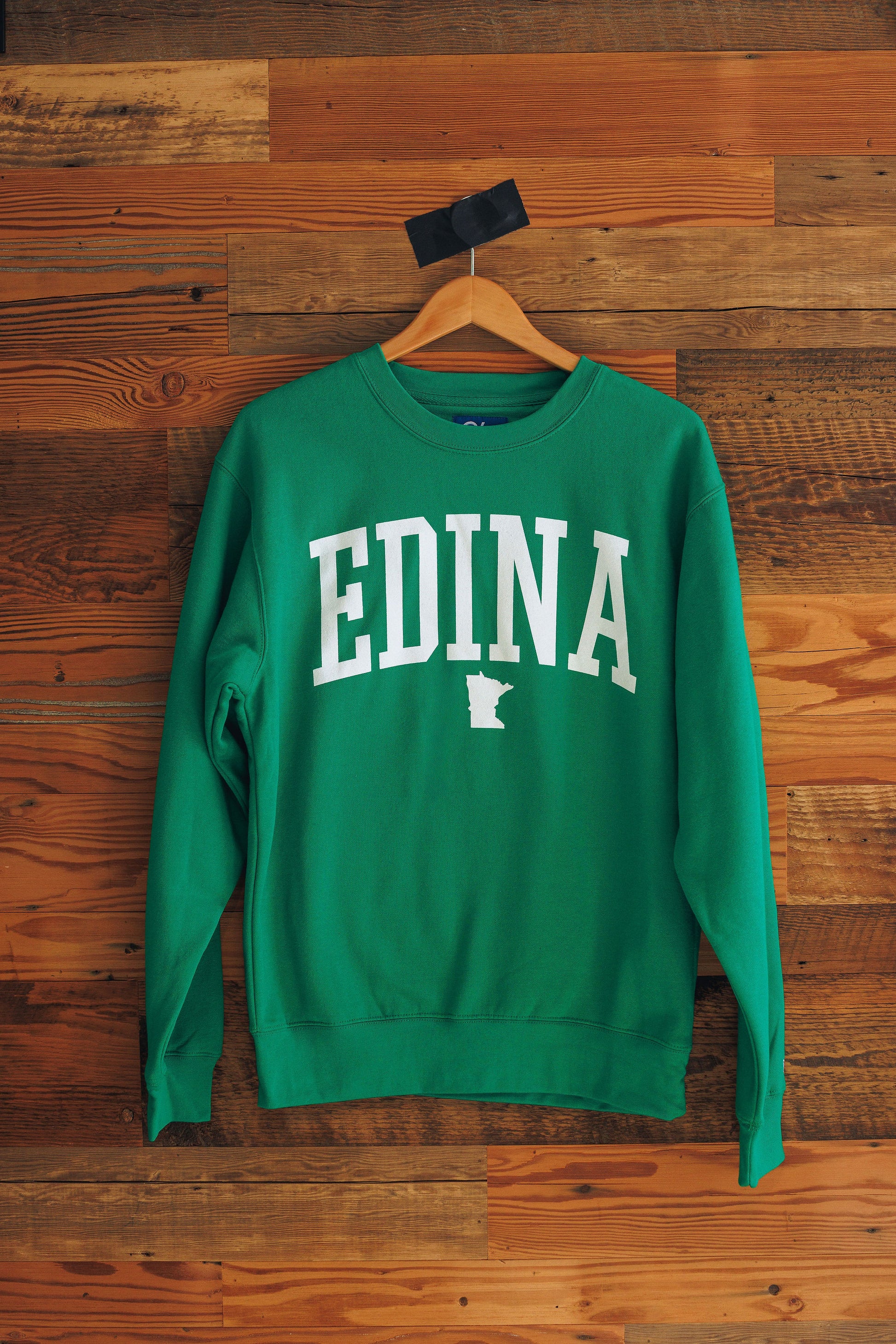 Green sweatshirt with 'Edina' printed on it, hanging against a wooden background