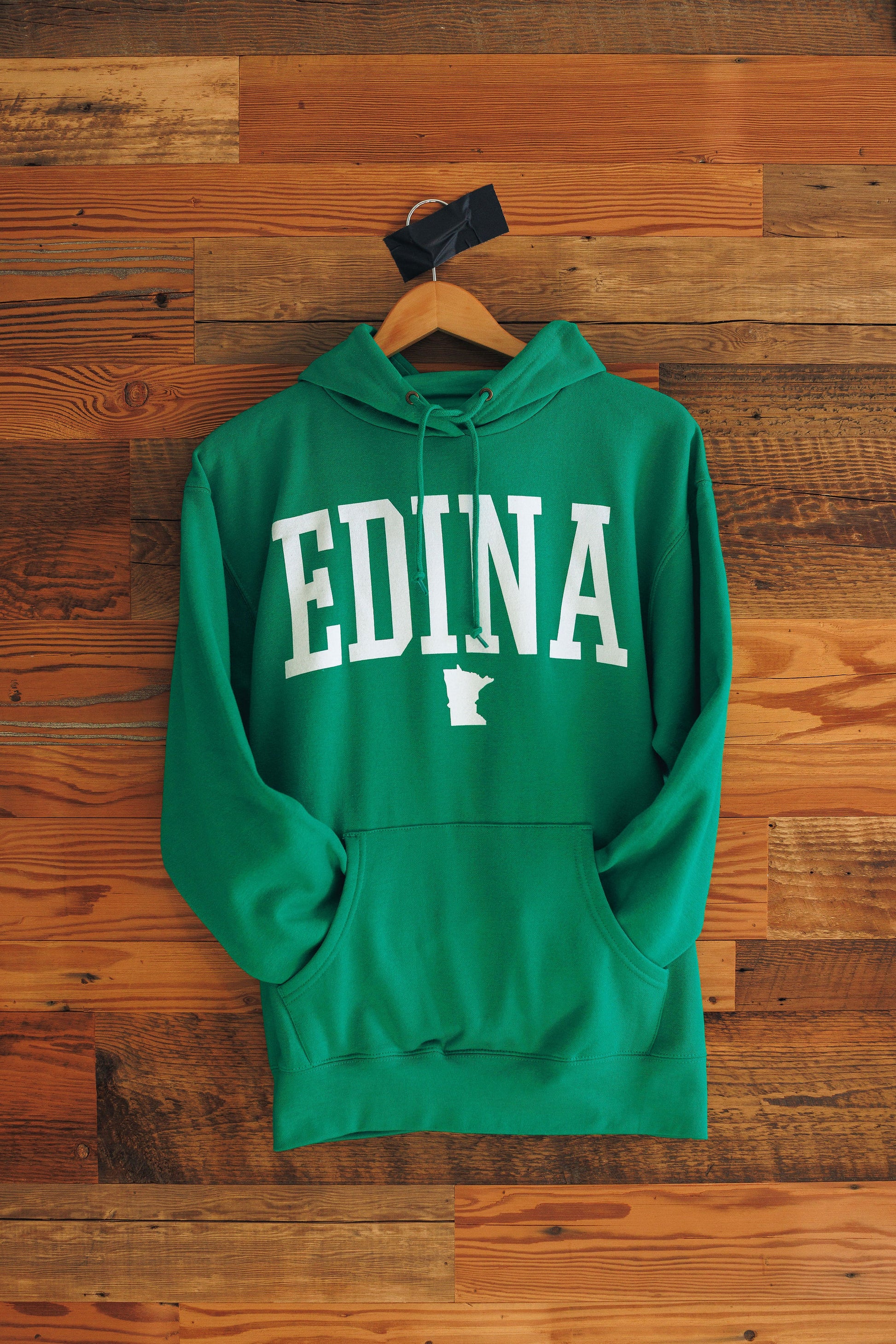 Green hoodie with 'EDINA' printed on it, hanging on a wooden hanger against a wooden background.