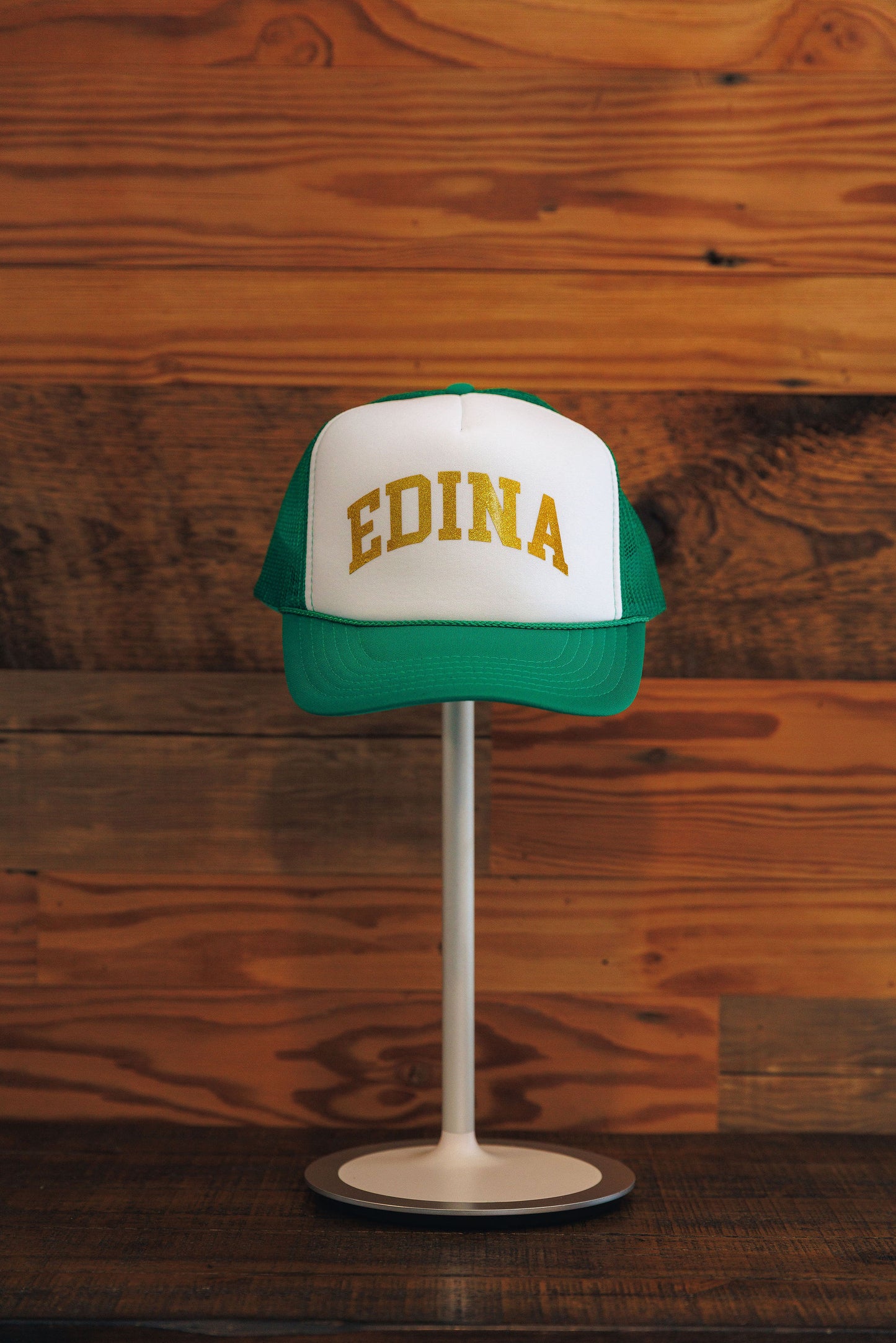 Green and white cap with 'Edina' text on a wooden stand against a wooden background