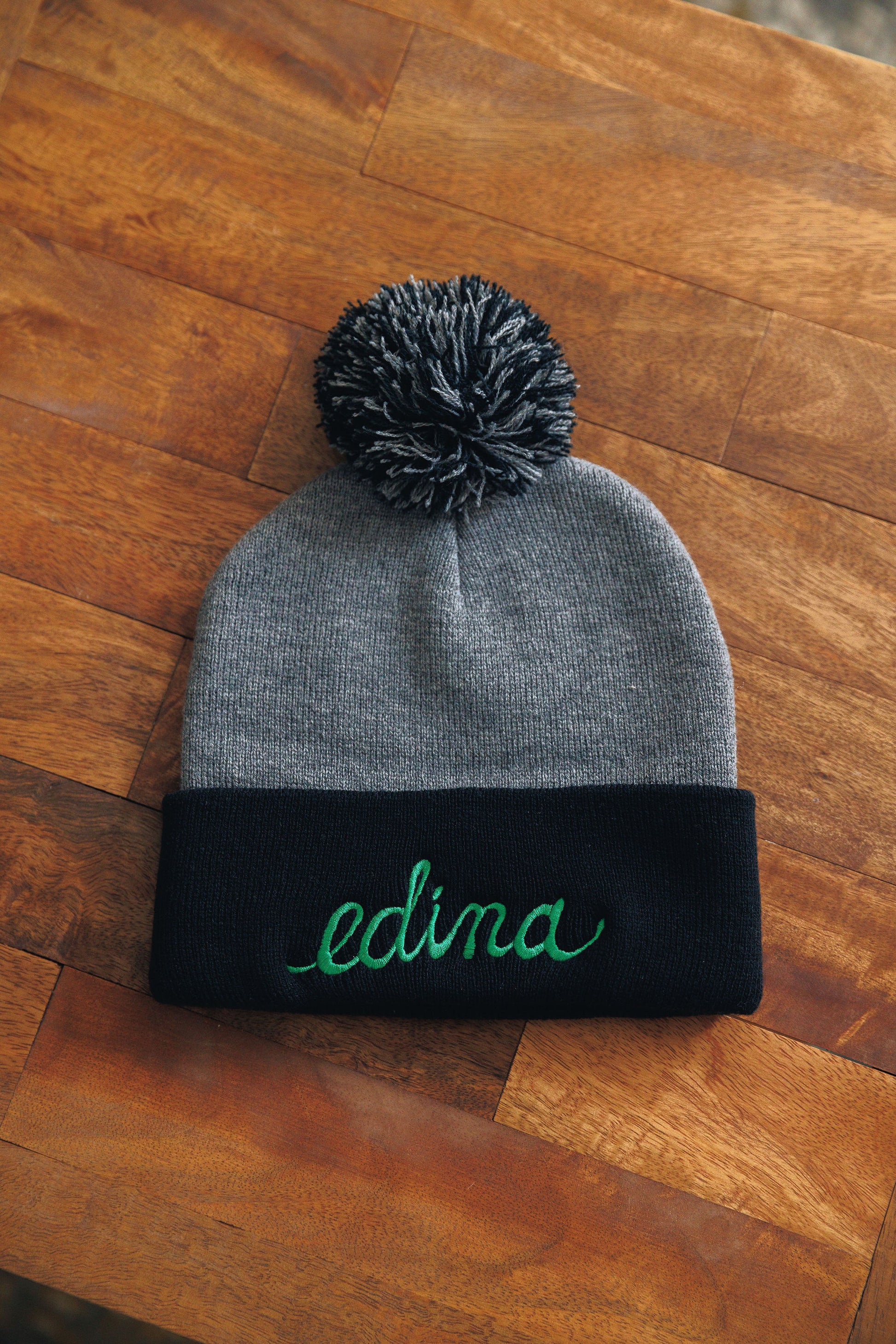 Gray beanie with black band and green 'edina' text on a wooden surface