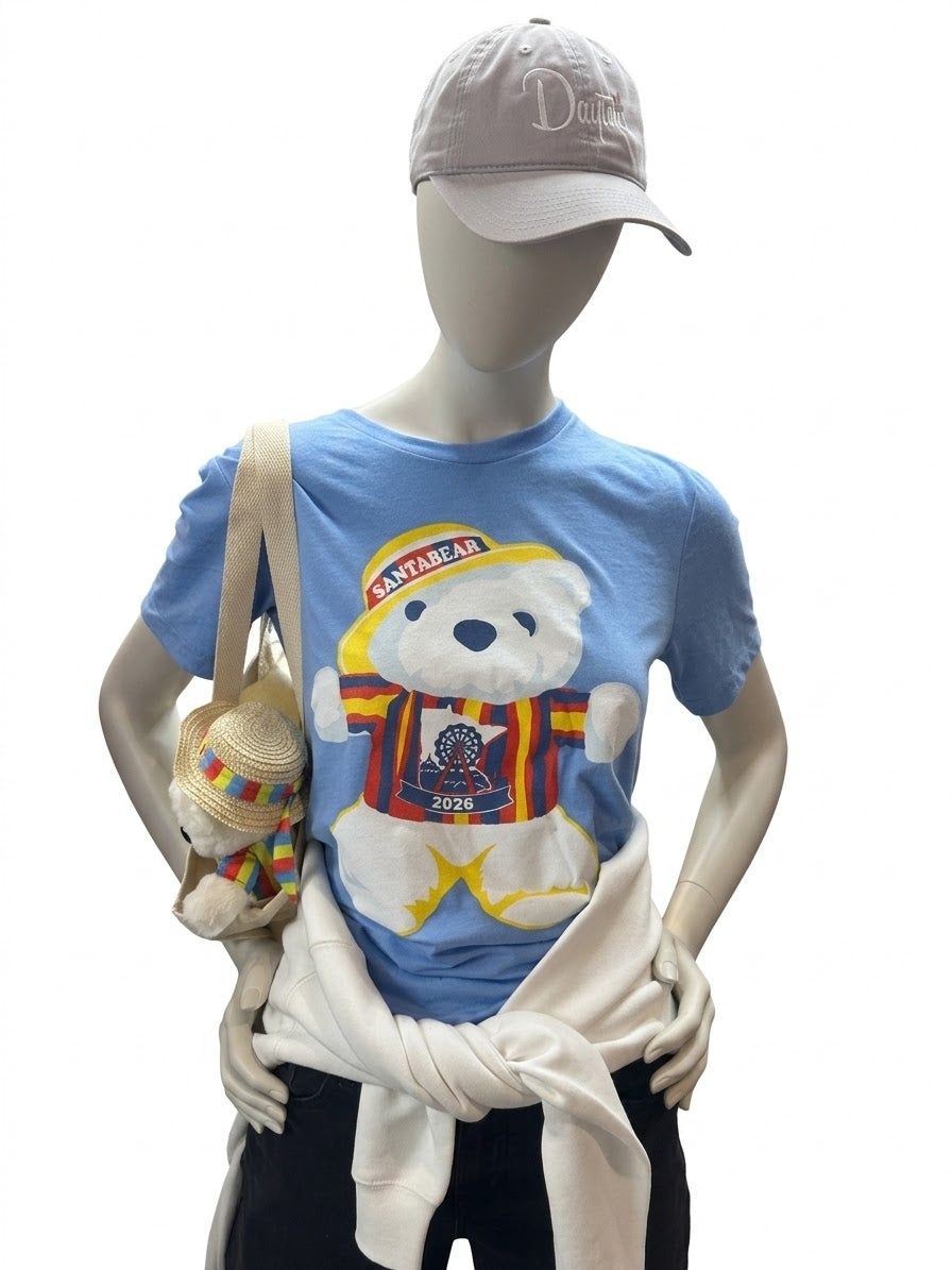A mannequin styled with a light blue T-shirt featuring a Summer Santabear who is wearing a yellow cap and striped jacket. Our mannequin has finished her look with a white sweatshirt tied around the waist, black pants, a gray Dayton's baseball cap, and a small woven shoulder bag.