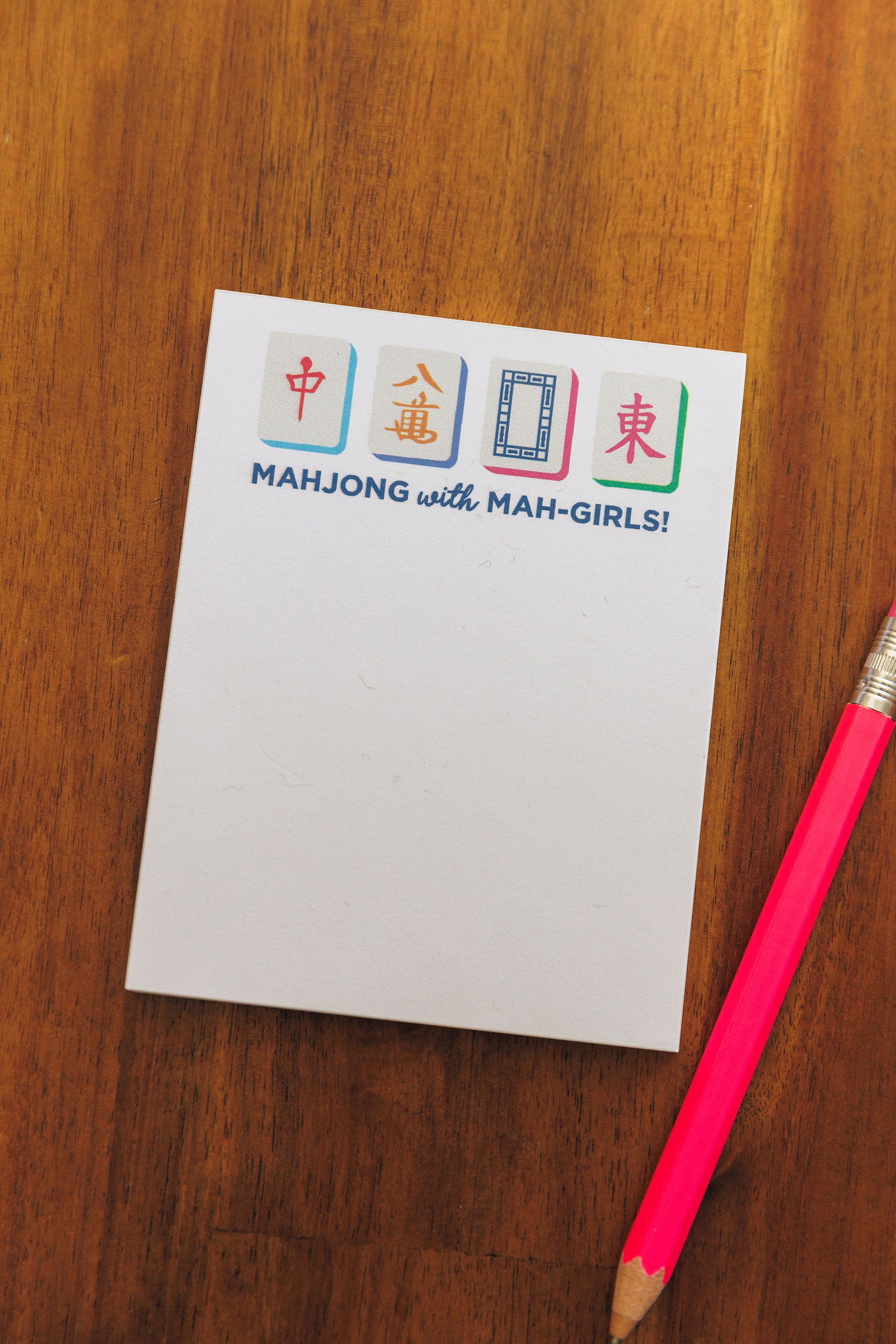 Notepad with 'Mahjong with Mah-Girls!' design on a wooden surface with a pink pencil.