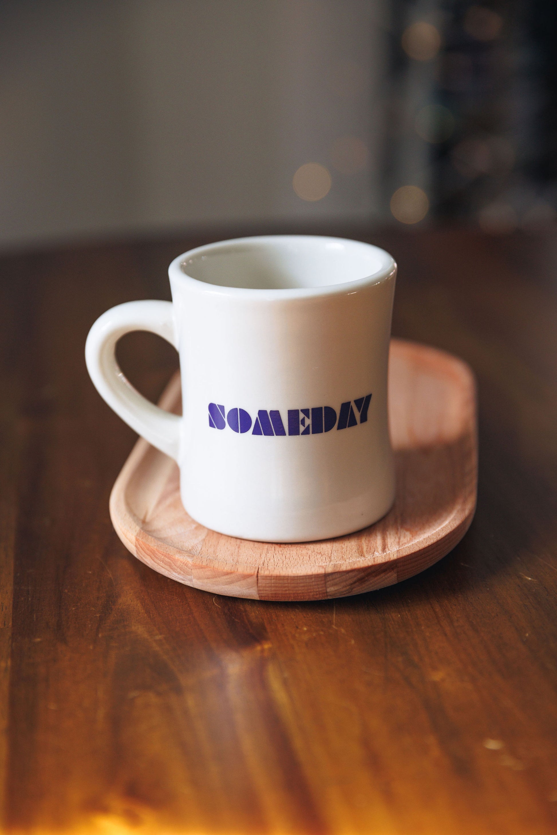 White mug with 'SOMEDAY' text on a wooden coaster on a wooden surface