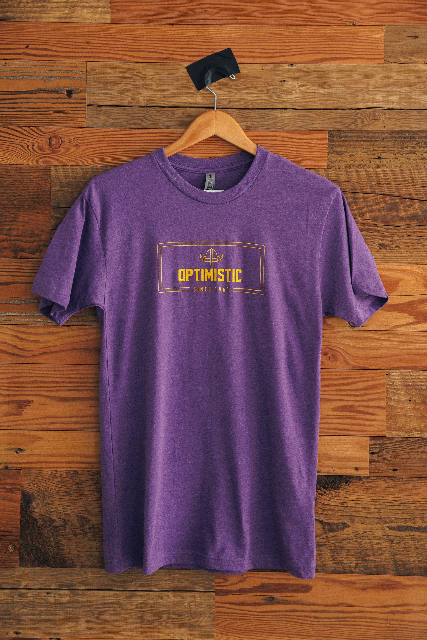 Purple t-shirt with 'Optimistic' text on a wooden wall