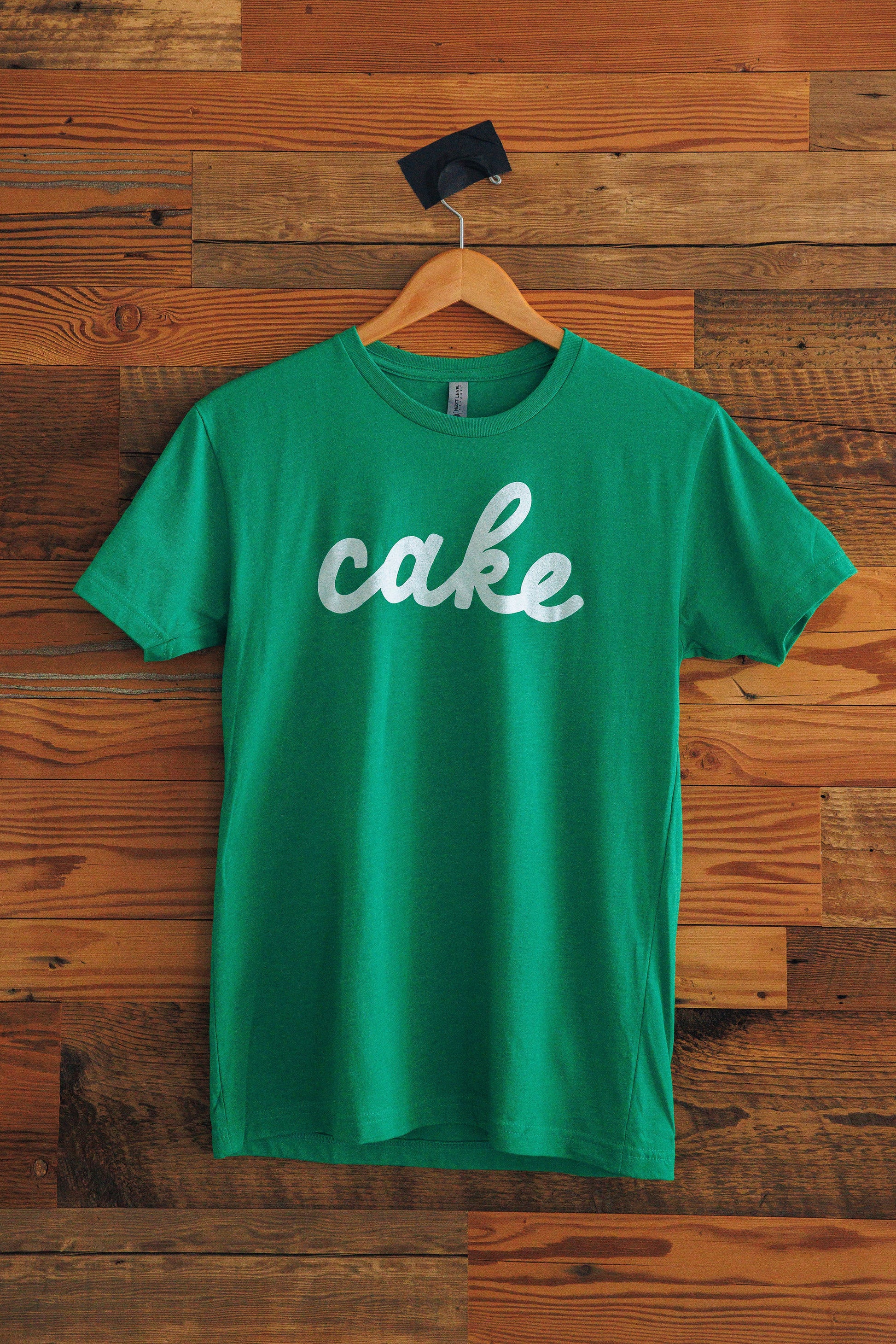Green t-shirt with 'cake' in cursive text on a wooden background
